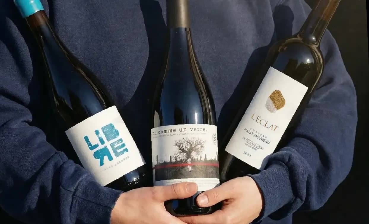 Person in blue shirt holding three distinct wine bottles with varied labels: 'Libre', 'Cheval de Bataille', 'L'Eclat'.