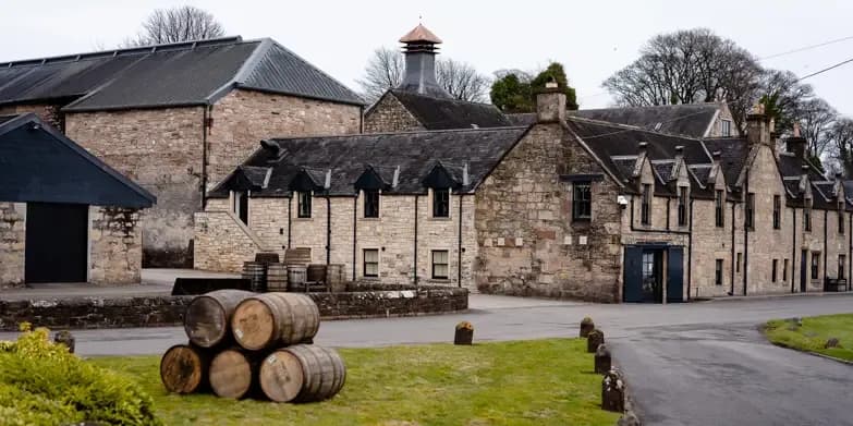 Old stone distillery buildings with stacked wooden barrels on green grass under a cloudy sky.