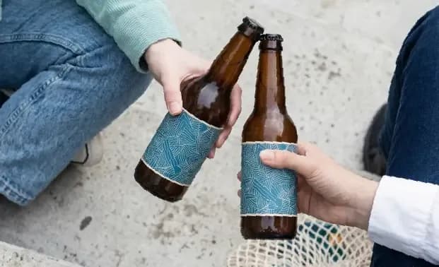 Two people clink brown beer bottles with patterned blue labels together in a toast.