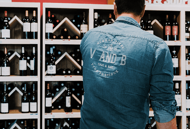 Man in denim shirt with V AND B text views wine bottles on shelves in a store.