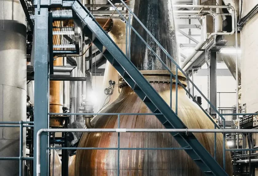 Large copper still and industrial stairs visible inside a bustling distillery facility.