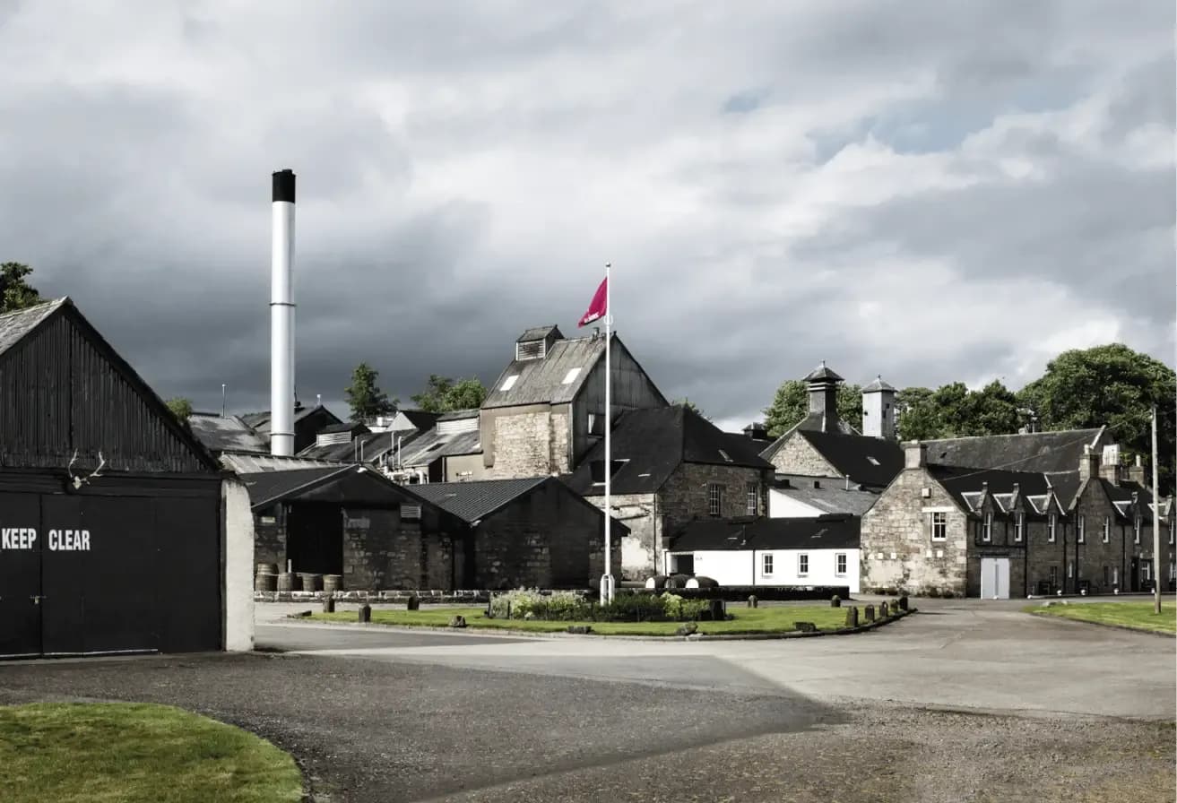 Historic distillery complex with dark buildings, a tall chimney, and a red flag under cloudy skies.