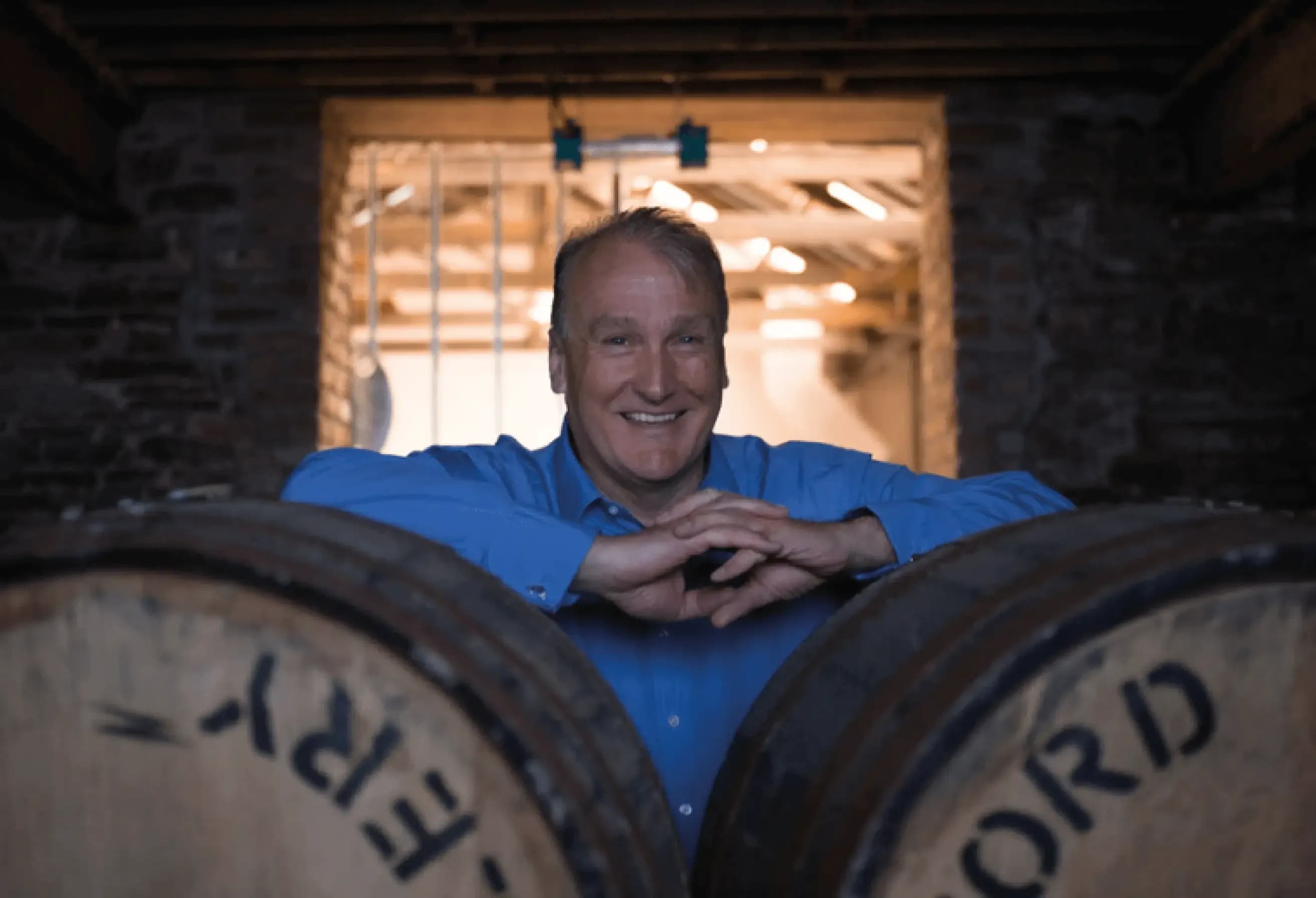 Smiling man in a blue shirt leaning on wooden whisky barrels inside a dimly lit distillery.