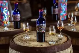 Colorful wine bottles with abstract labels and glasses with amber liquid displayed on wooden barrels.