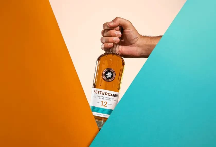 A hand holds a bottle of Piet Heijn 12 rum against a split orange and teal background.