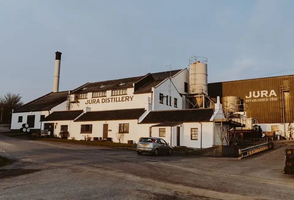 White Jura Distillery building with black roofs, a tall chimney, and parked car.