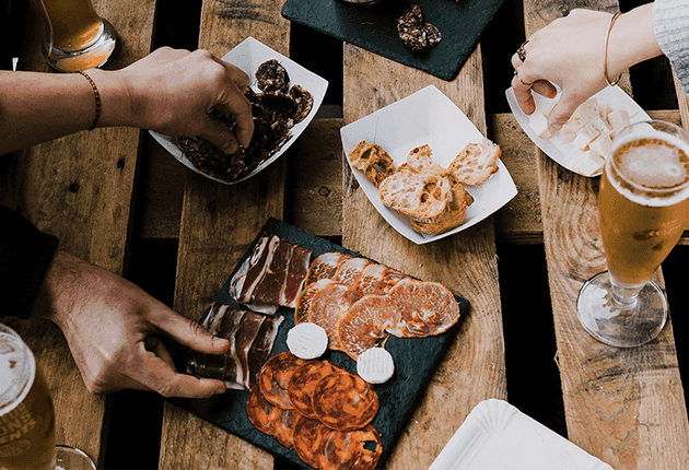 Hands reach for an assortment of cured meats, cheeses, and snacks on a rustic wooden table.