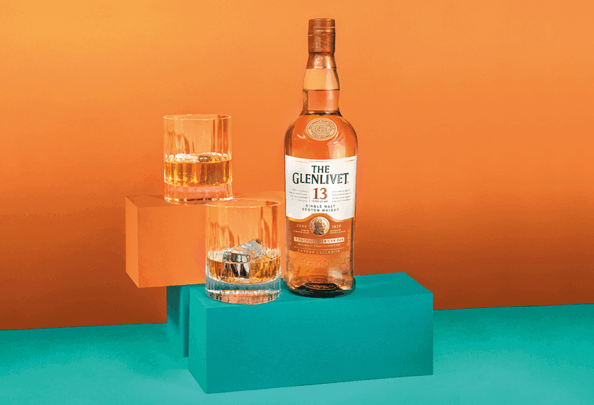 Glenlivet 13 whiskey bottle and two glasses, one with ice, on colorful risers.