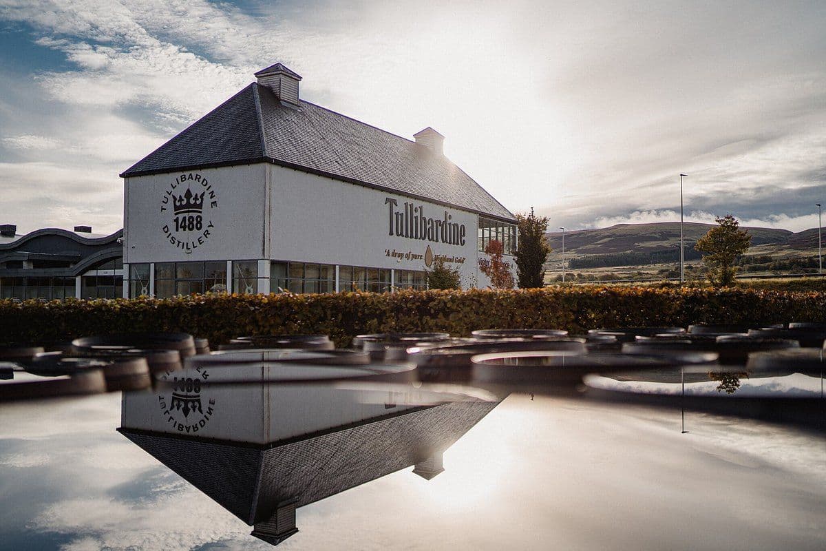 Tullibardine distillery building reflected in water with barrels under a bright sky.