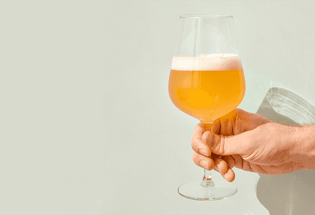 A hand holds a stemmed glass filled with frothy golden beer against a plain background.