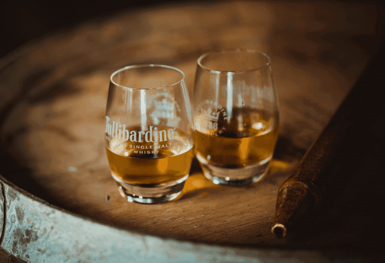Two whiskey glasses with amber liquid and a cigar rest on a rustic wooden barrel.