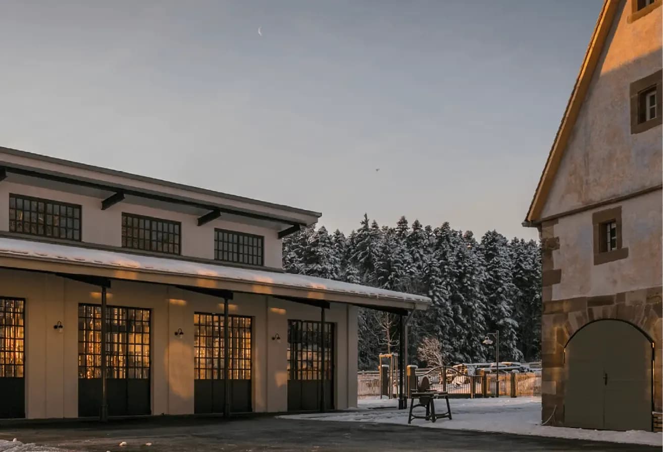 Two snow-covered buildings with warm light reflecting off windows, backed by a winter forest.